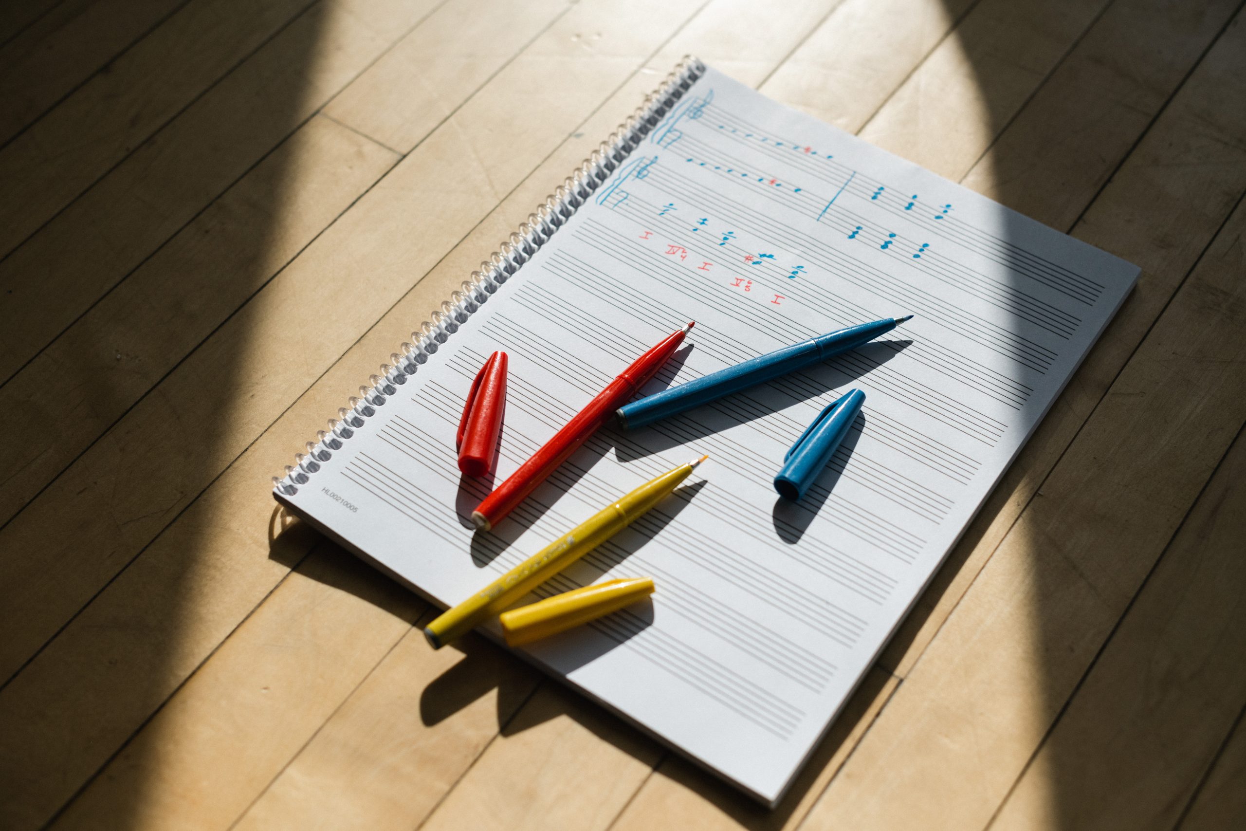 An image of a notebook containing music staff and on top are colorful pens and pencils